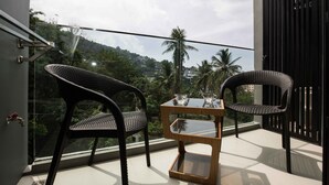A balcony with two wicker chairs, a glass-top table, and a view of a green landscape with palm trees and buildings. - The Panora apartments by NLA (Choeng Thale)