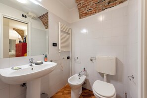 Un baño con lavamanos, bidé y retrete blancos, suelo de madera y pared de ladrillo.