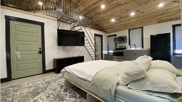 Une chambre à coucher comprenant un lit, un téléviseur fixé au mur, un escalier et un coin cuisine équipé d’appareils électroménagers.