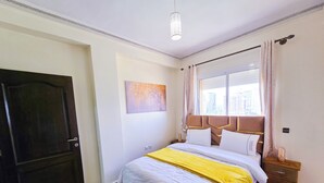 Ein Schlafzimmer mit Bett, Nachttisch, Fenster mit Vorhängen, einer Tür und einem Wandbild.
