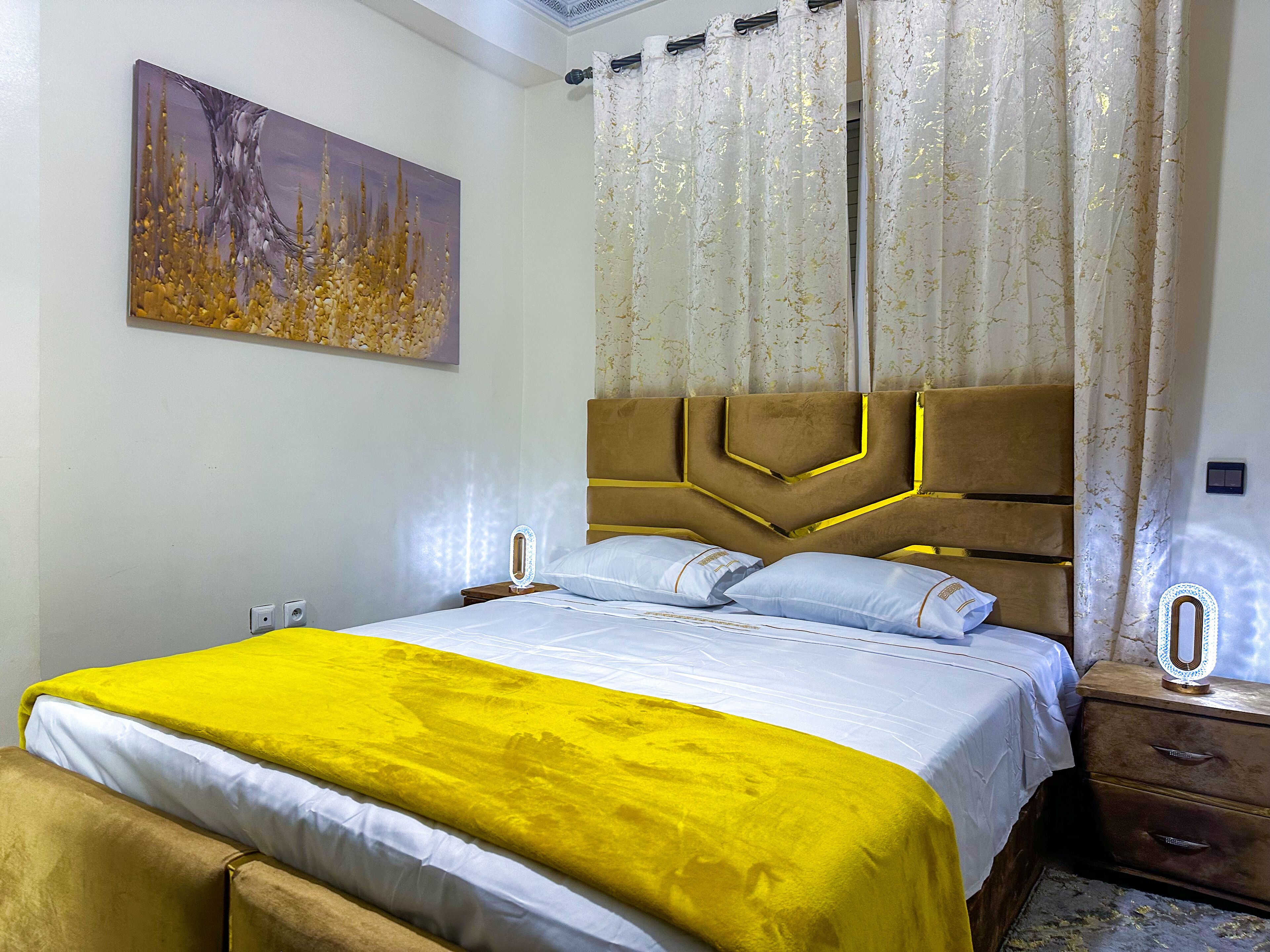A neatly made bed with a yellow bedspread, a headboard with geometric patterns, and a painting on the wall.
