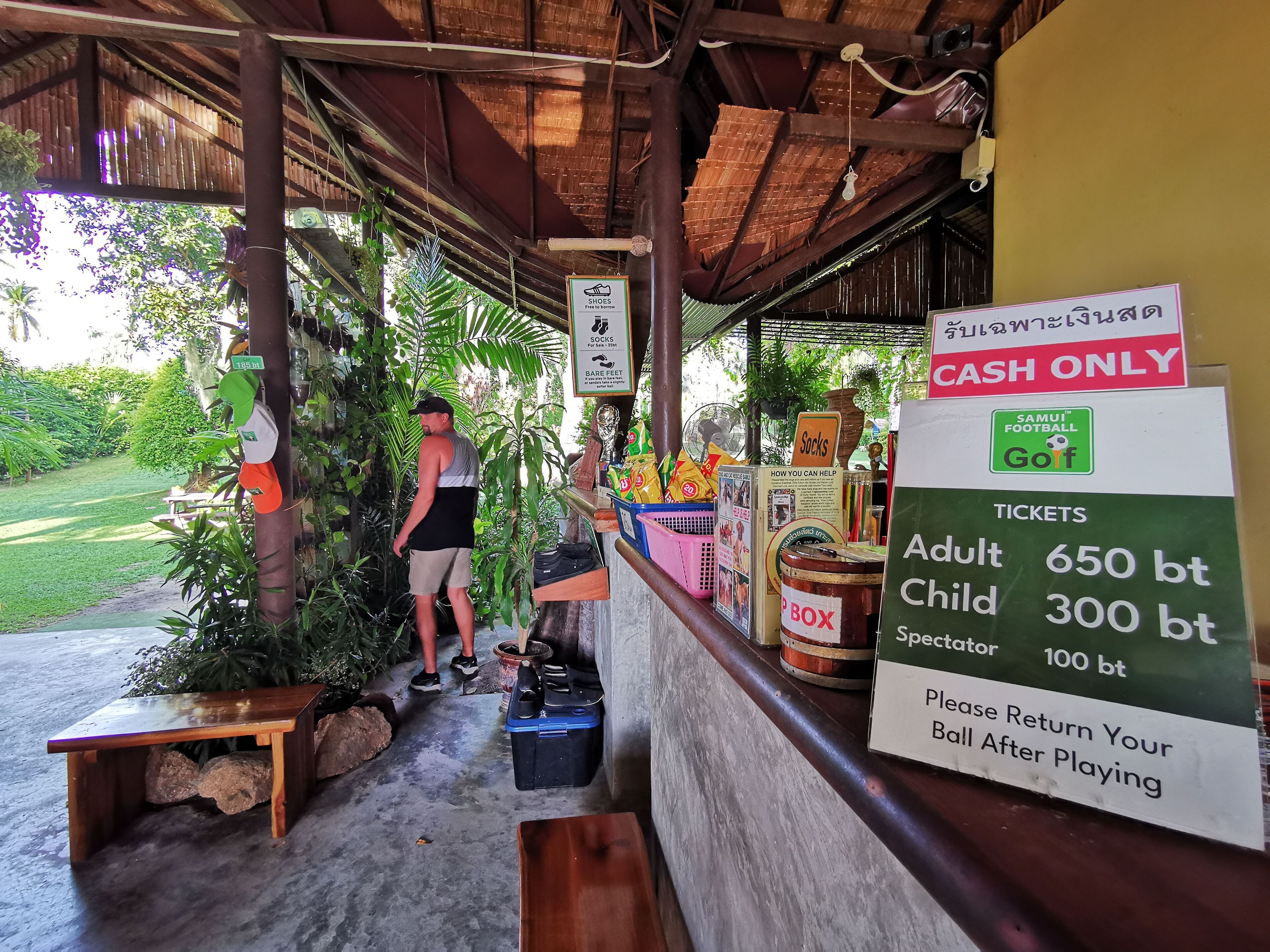 A covered outdoor area with a wooden bench, a table, and a sign indicating ticket prices for adults, children, and spectators.