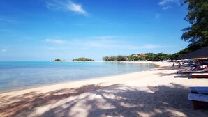 Una spiaggia con acque cristalline, una riva sabbiosa e una piccola isola all'orizzonte. Sulla spiaggia si vedono lettini e ombrelloni.