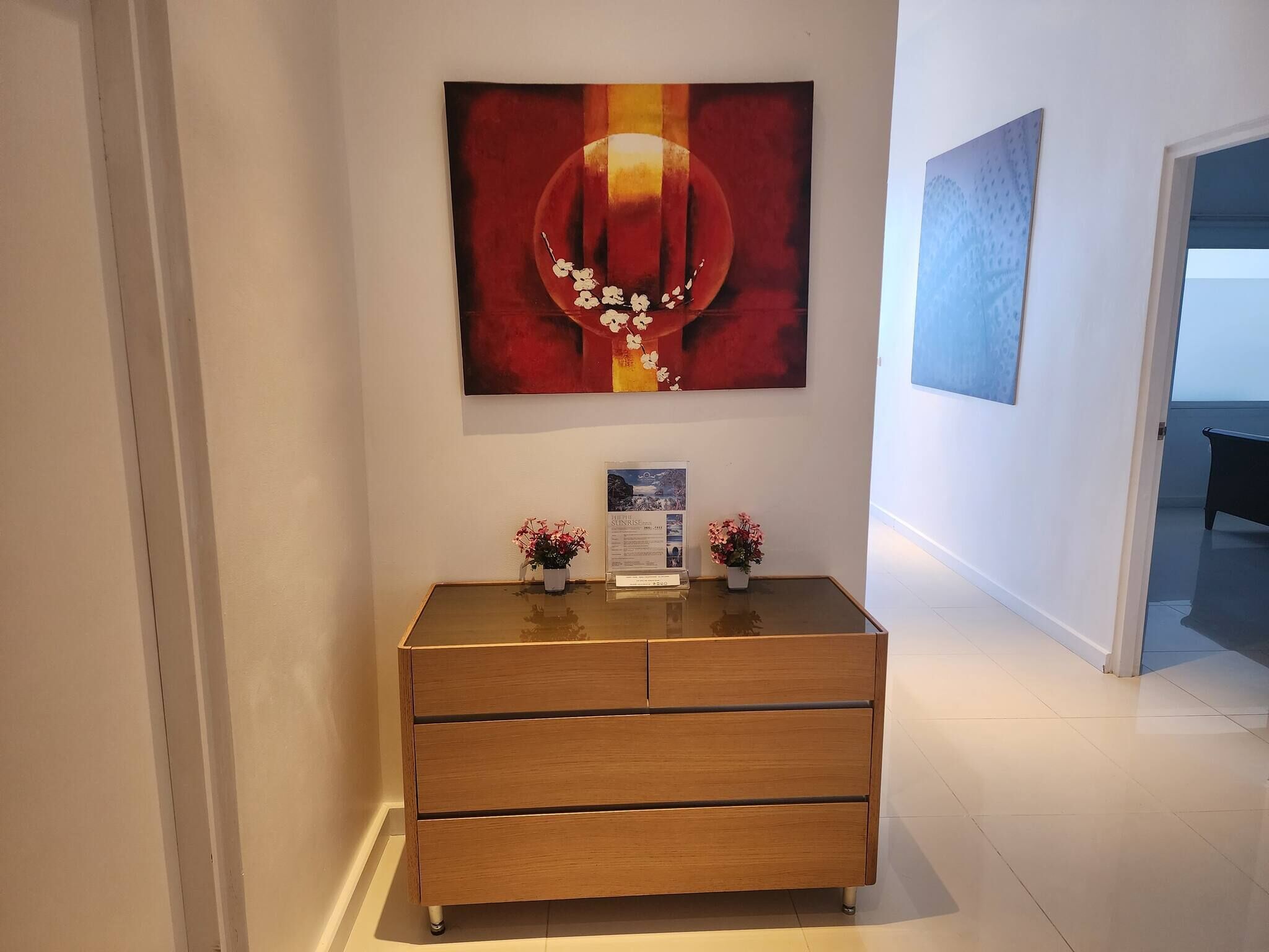 Un couloir avec une commode en bois, une œuvre d’art encadrée et une petite table ornée de fleurs.