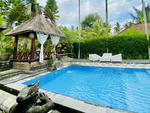 Una piscina con un quiosco de techo de paja, sillas reclinables blancas y una estatua.