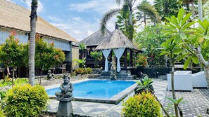 Una piscina rodeada de plantas tropicales, una estatua y una estructura con techo de paja.