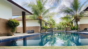Zona de piscina con sillas de descanso, palmeras y una piscina azul cristalina.