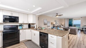 A modern kitchen with white cabinets, a black microwave, and a dishwasher. There is a dining area with a gray couch and a view of the ocean through large windows.