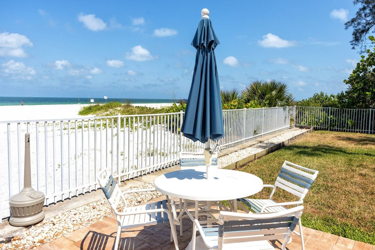 A patio with a white table and chairs, a blue umbrella, and a view of the beach.