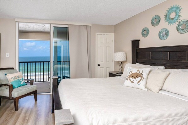 A bedroom with a large bed, a chair, a nightstand, a lamp, and a view of the ocean through a sliding door.