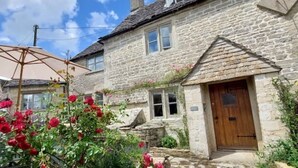 A stone house with a wooden door, a garden with red flowers, and a patio umbrella. - Luxury Cosy Cottage In Magical Bibury (Bibury)