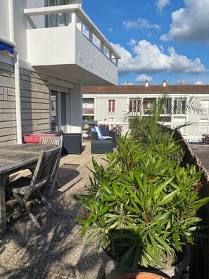 Ein Außenbereich mit einer Holzterrasse, einem Tisch und Stühlen, einem Blumentopf und Blick auf ein Gebäude mit Balkonen und Fenstern.