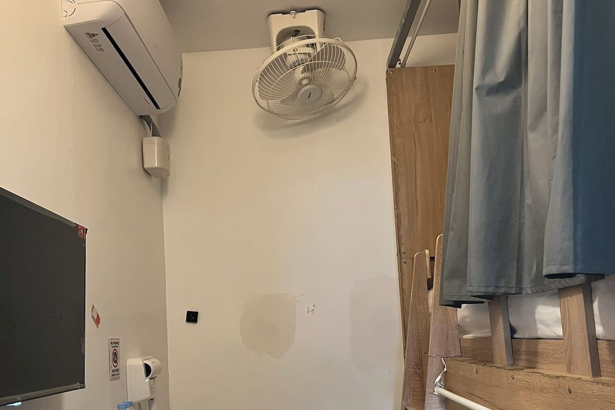 A compact room with a bed, a small stool, a shelf with a bottle and a remote, an air conditioner, and a ceiling fan.