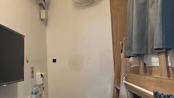 Ein kleines Zimmer mit einem Bett, einem kleinen Hocker, einem Regal mit einer Flasche und einer Fernbedienung, einer Klimaanlage und einem Deckenventilator.