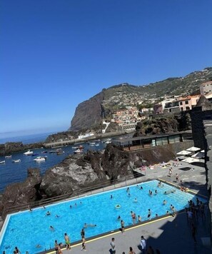 Ein Poolbereich mit Schwimmern und Sonnenanbetern, mit Blick auf eine felsige Küstenlinie mit Gebäuden und Booten.
