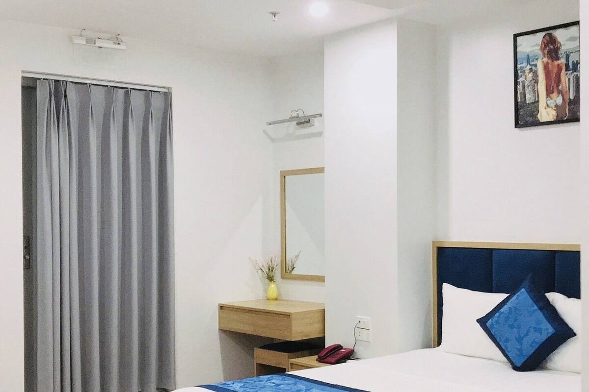 A hotel room with a bed, a bedside table, a mirror, a window with curtains, and a ceiling fan.