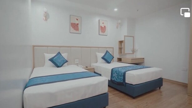 Une chambre d’hôtel avec deux lits, chacun doté d’une tête de lit bleue et d’un oreiller assorti. Une table de chevet en bois se trouve entre les deux lits, ainsi qu’une petite table en bois sur laquelle reposent un miroir et une étagère.