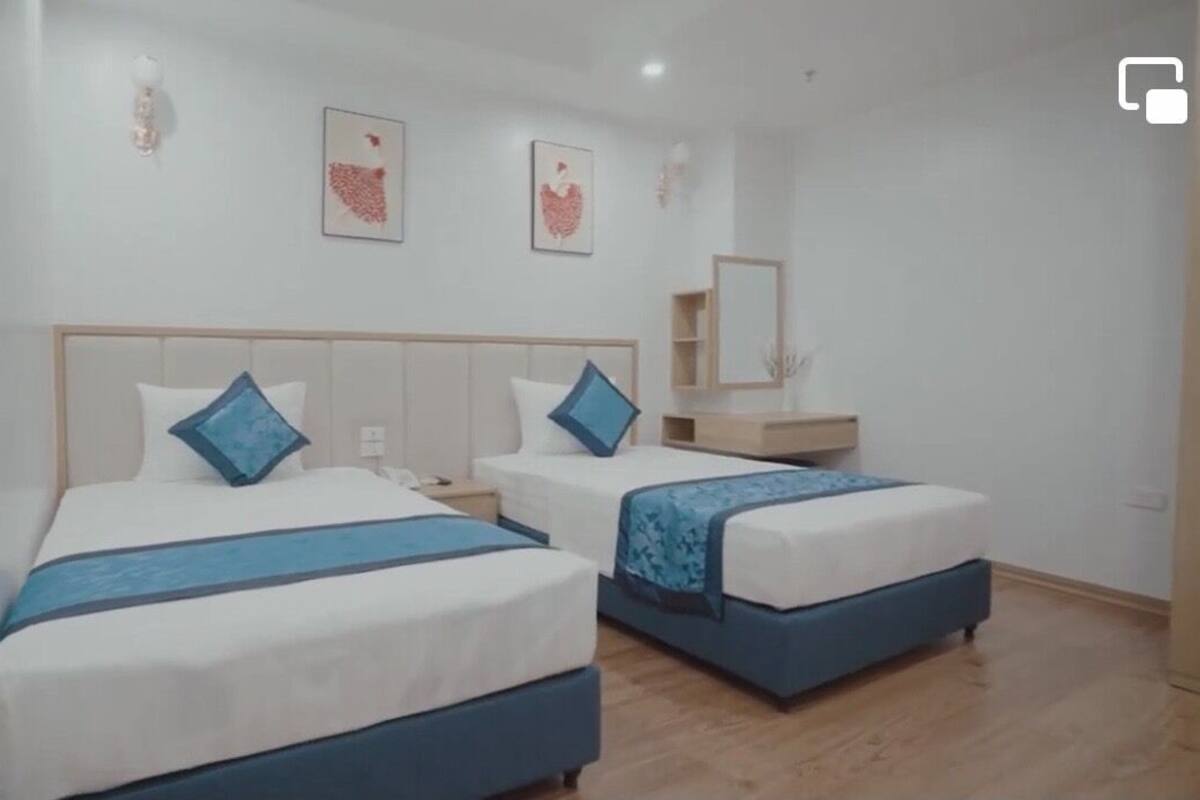 A hotel room with two beds, each with a blue headboard and matching pillow. There is a wooden nightstand between the beds, and a small wooden table with a mirror and a shelf above it.