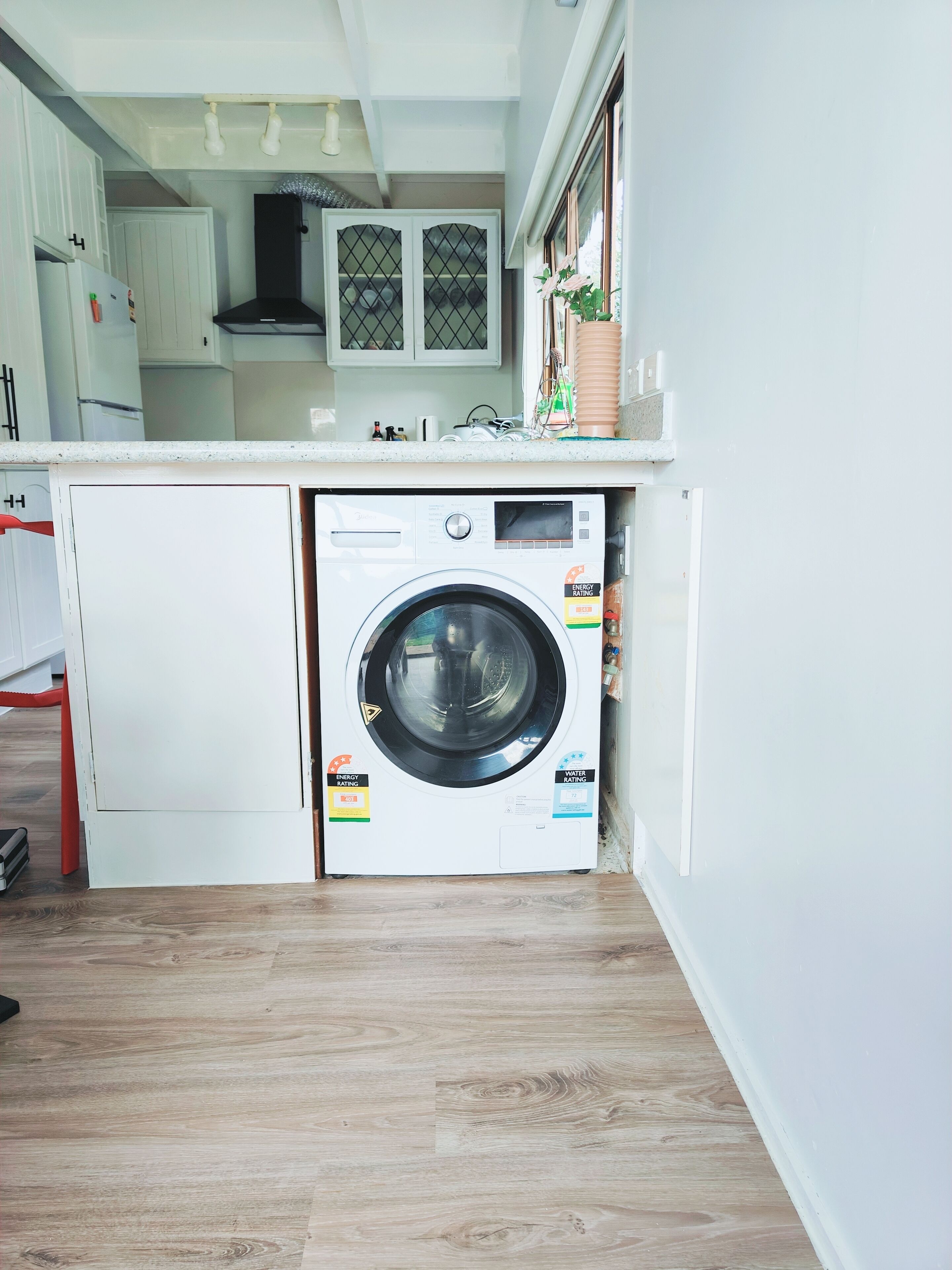 Une cuisine compacte dotée d’un lave-linge et d’un sèche-linge intégrés, d’un plancher en bois et de meubles de cuisine blancs.