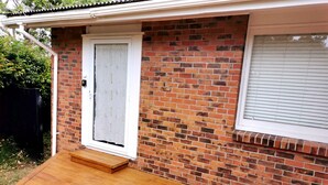 Une maison en briques avec une terrasse en bois, une porte blanche et une fenêtre avec des stores.