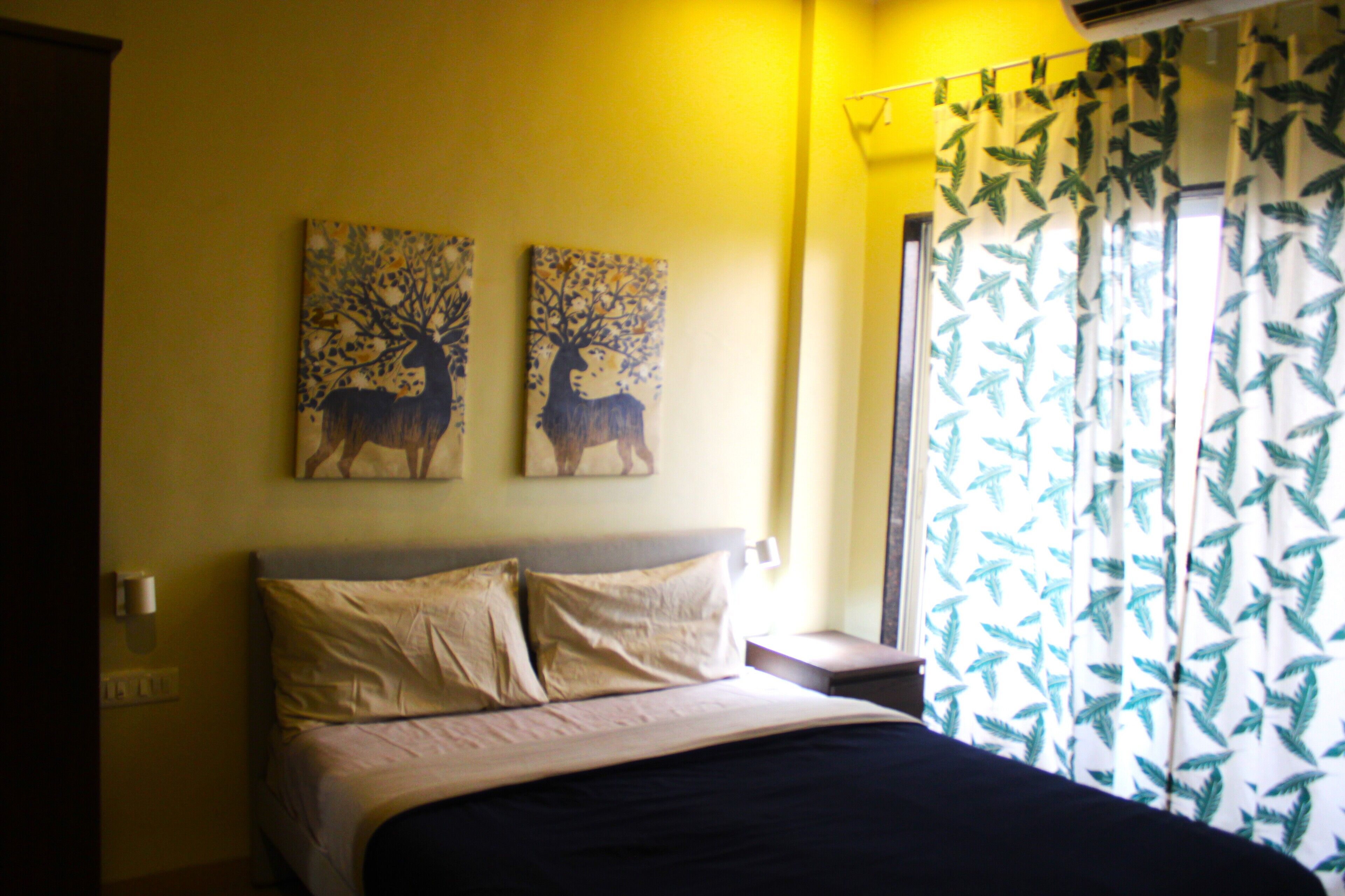 A bedroom with a bed, two wall-mounted paintings of deer, a nightstand, and a window with leaf-patterned curtains.