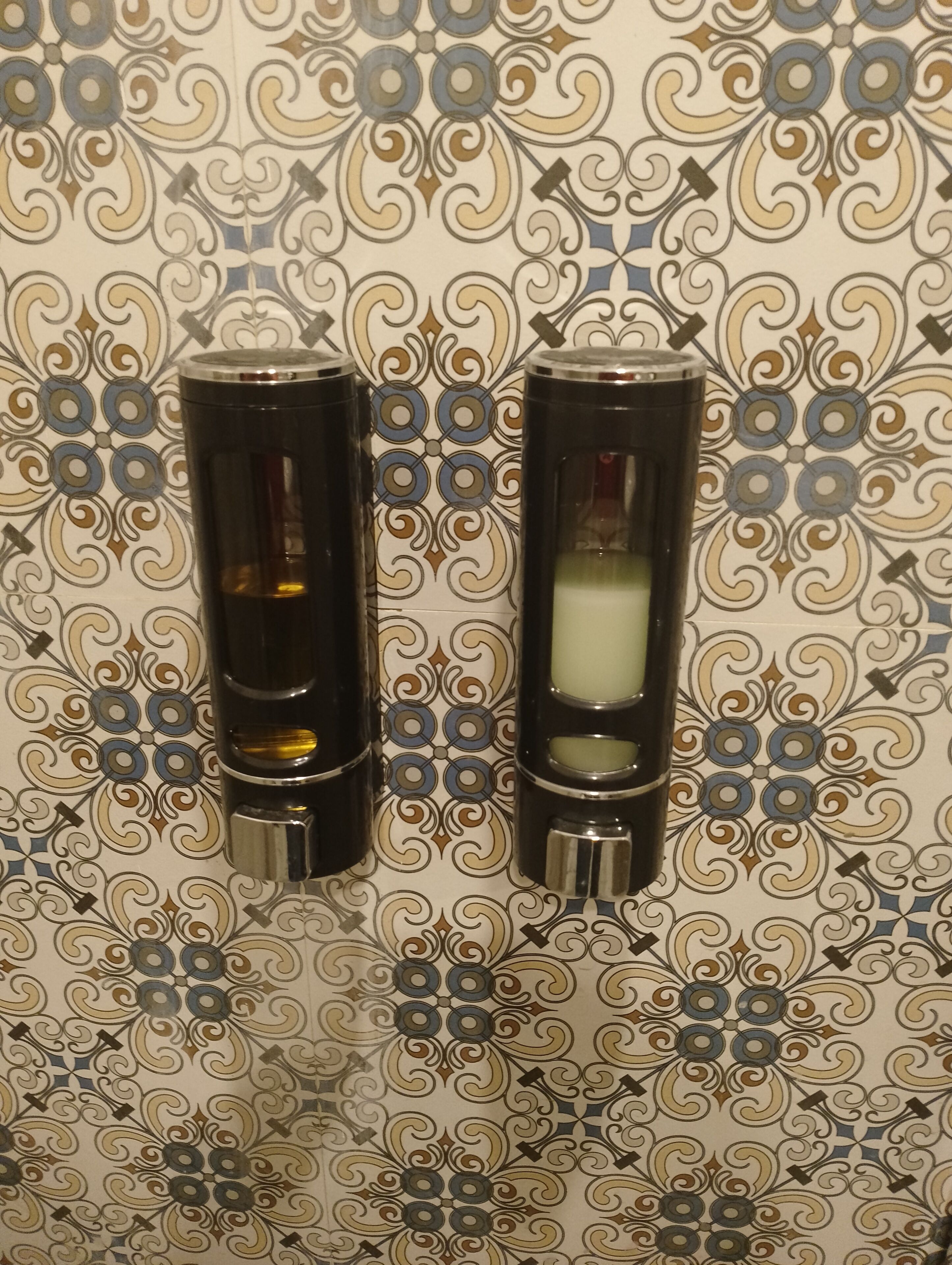 Two black dispensers with a transparent section, mounted on a wall with a decorative tile pattern.