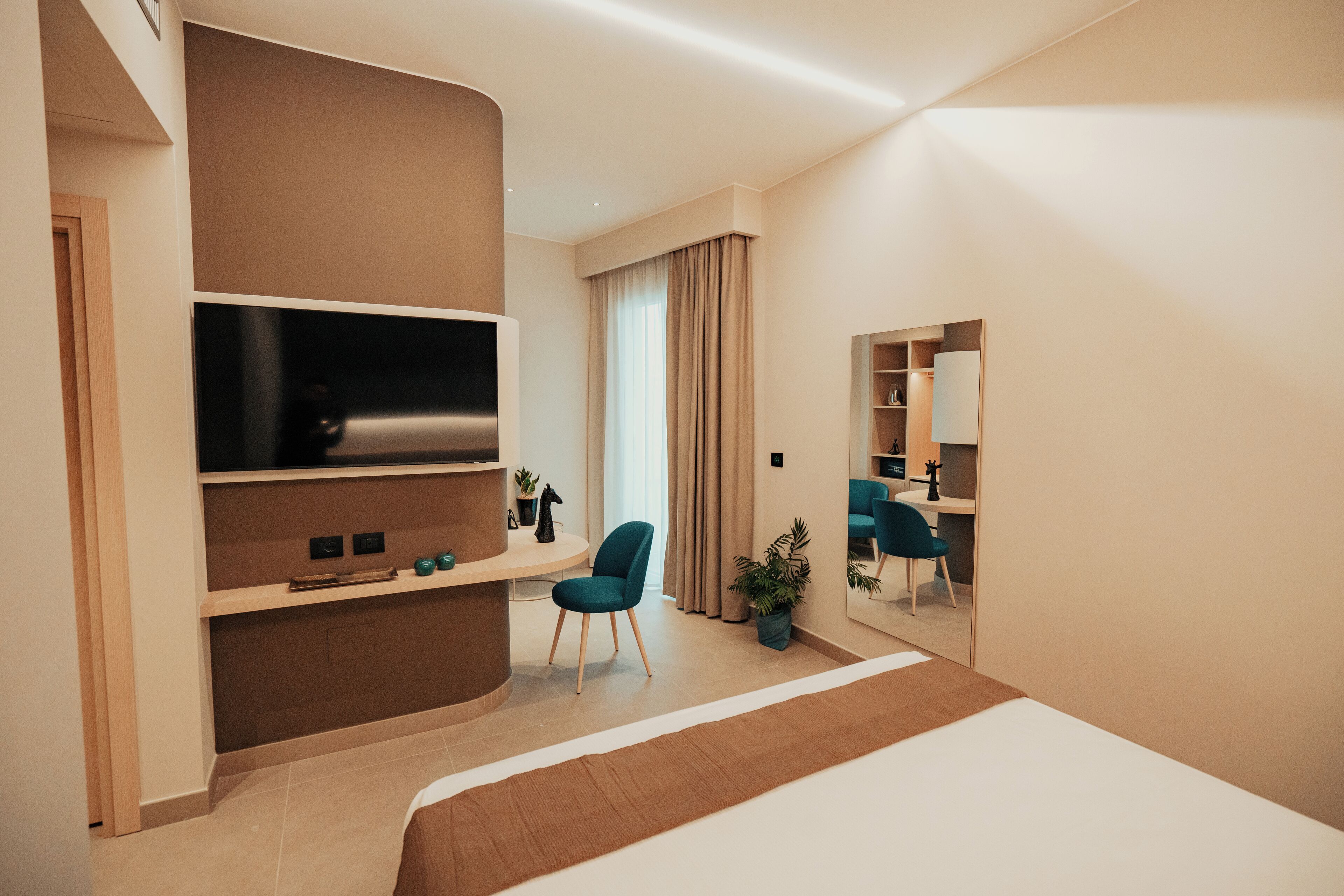 A modern hotel room with a large bed, a flat-screen TV mounted on a wall, a desk with a chair, and a mirror reflecting the room.