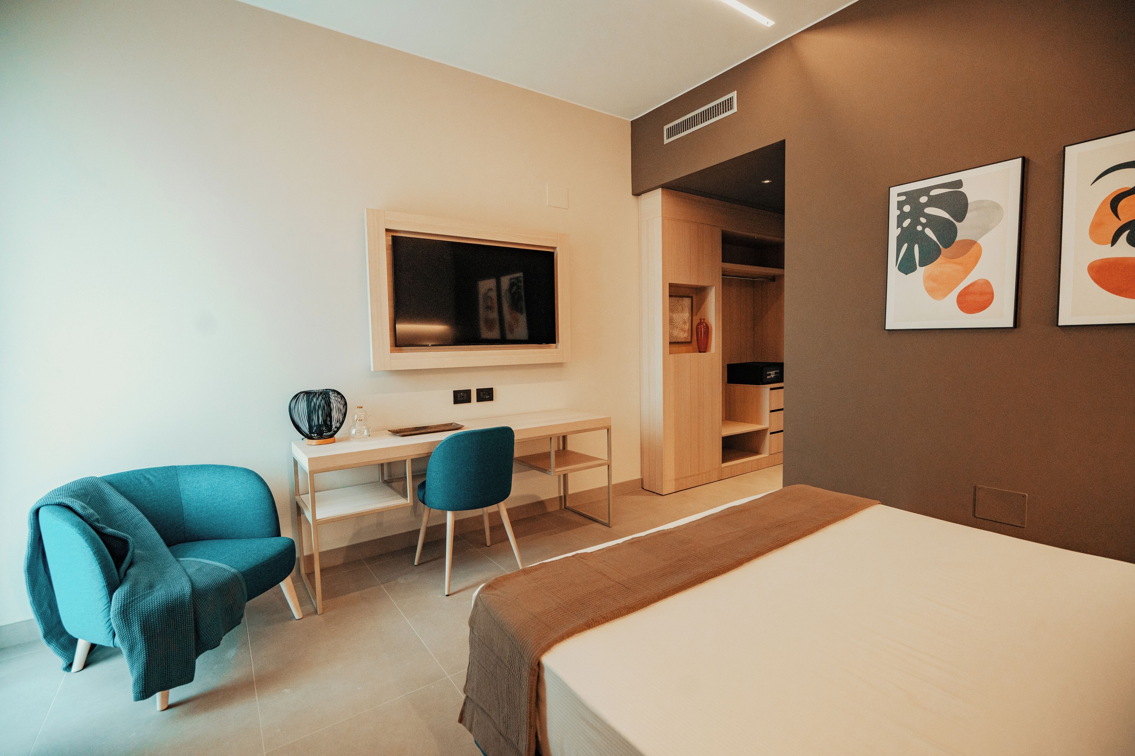 A modern hotel room with a bed, a desk, two chairs, a wardrobe, and artwork on the walls.