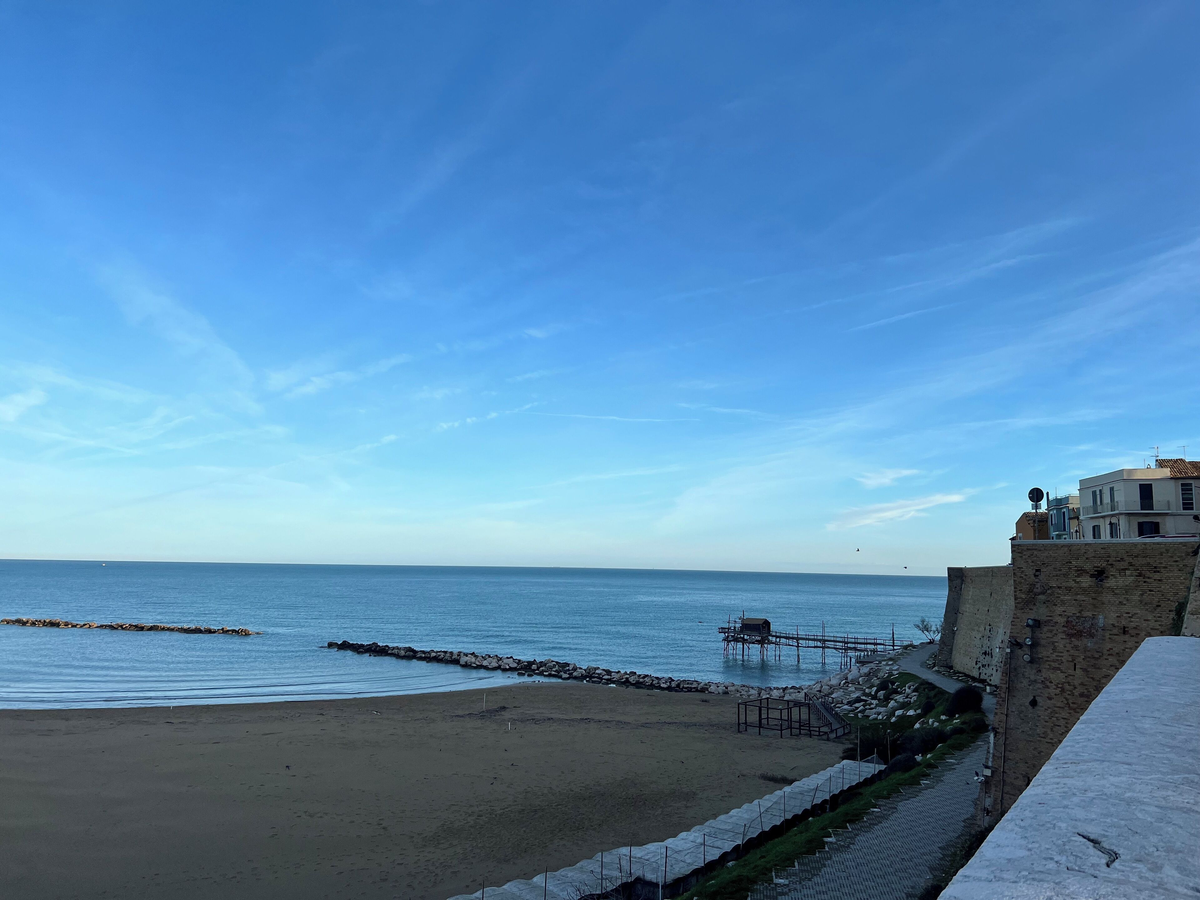 A coastal view with a sandy beach, a stone wall, and a building with a balcony overlooking the sea.
