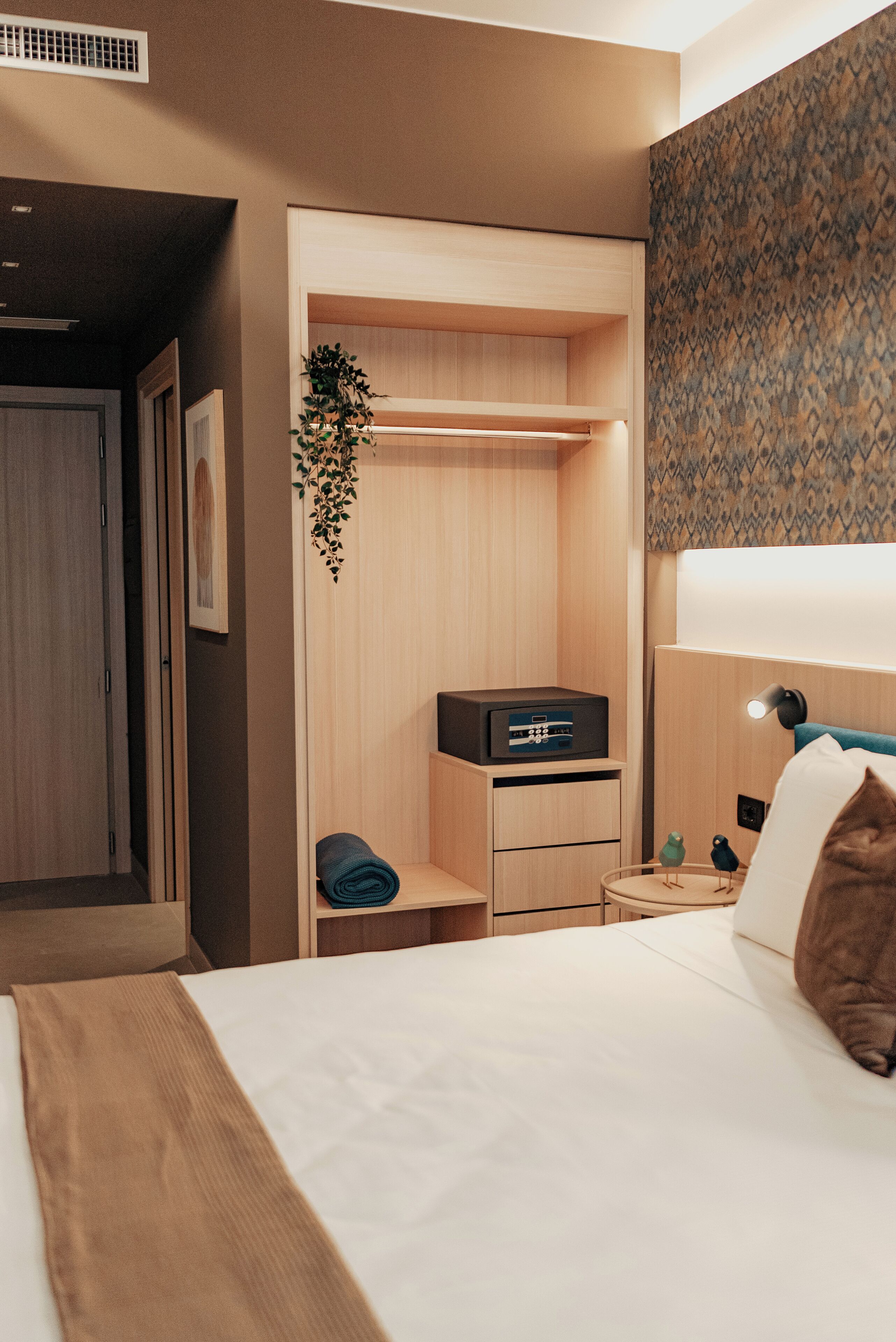 A hotel room with a bed, a desk with a lamp, a small plant, and a wall-mounted shelf.