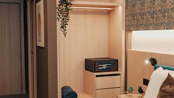 A hotel room with a bed, a desk with a lamp, a small plant, and a wall-mounted shelf.