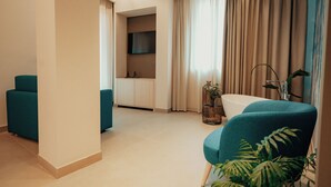 A modern hotel room with teal armchairs, a white bathtub, a sink, and a flat-screen TV mounted on the wall.
