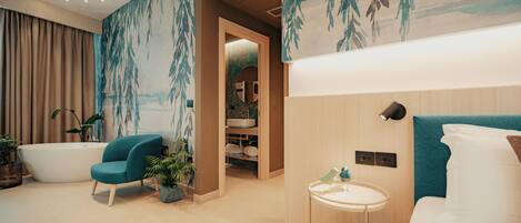 A modern bathroom with a bathtub, a sink, and a shower area. There is a chair and a small table with a lamp.
