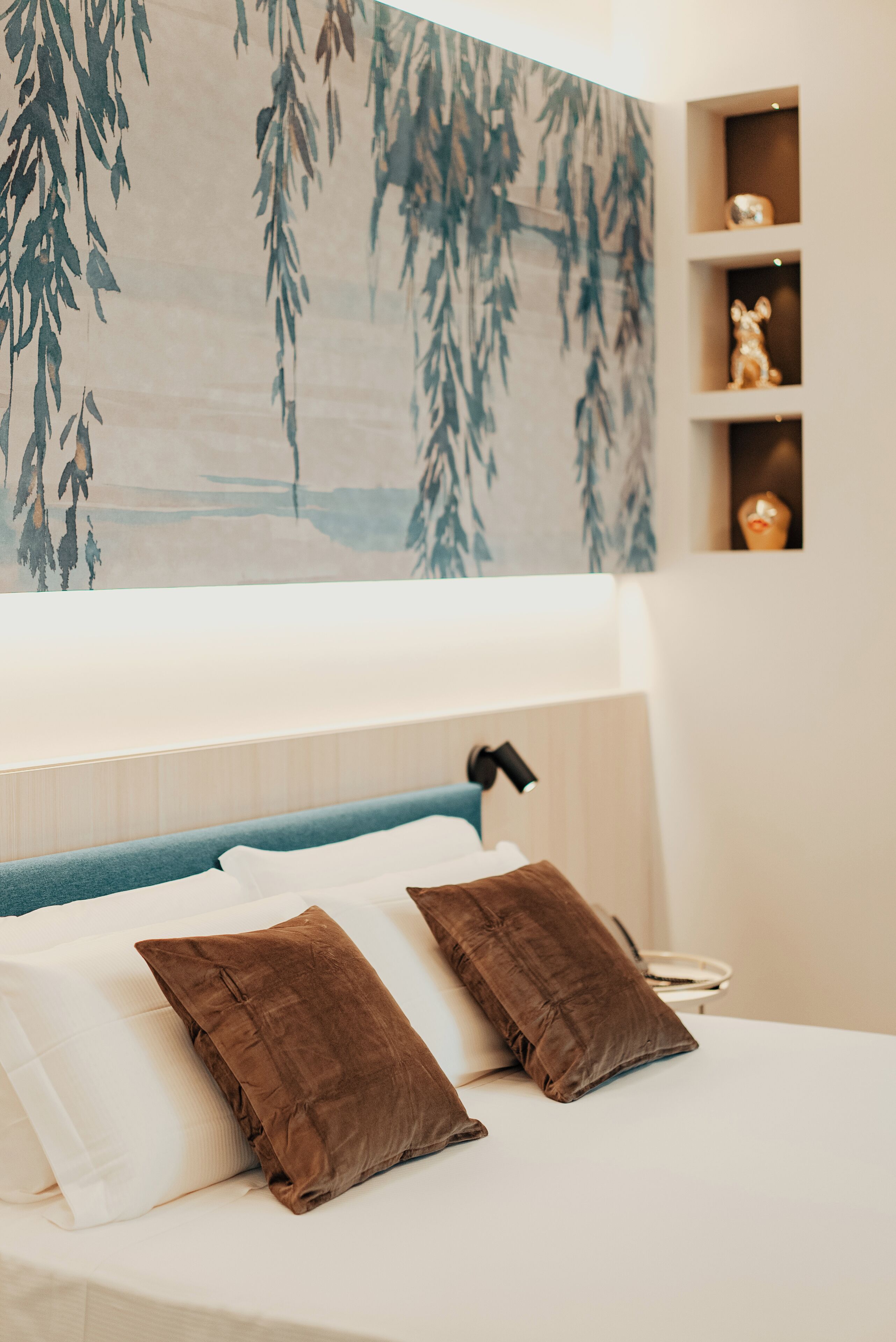 A bedroom with a bed, pillows, a nightstand, a lamp, and a wall with a hanging plant.