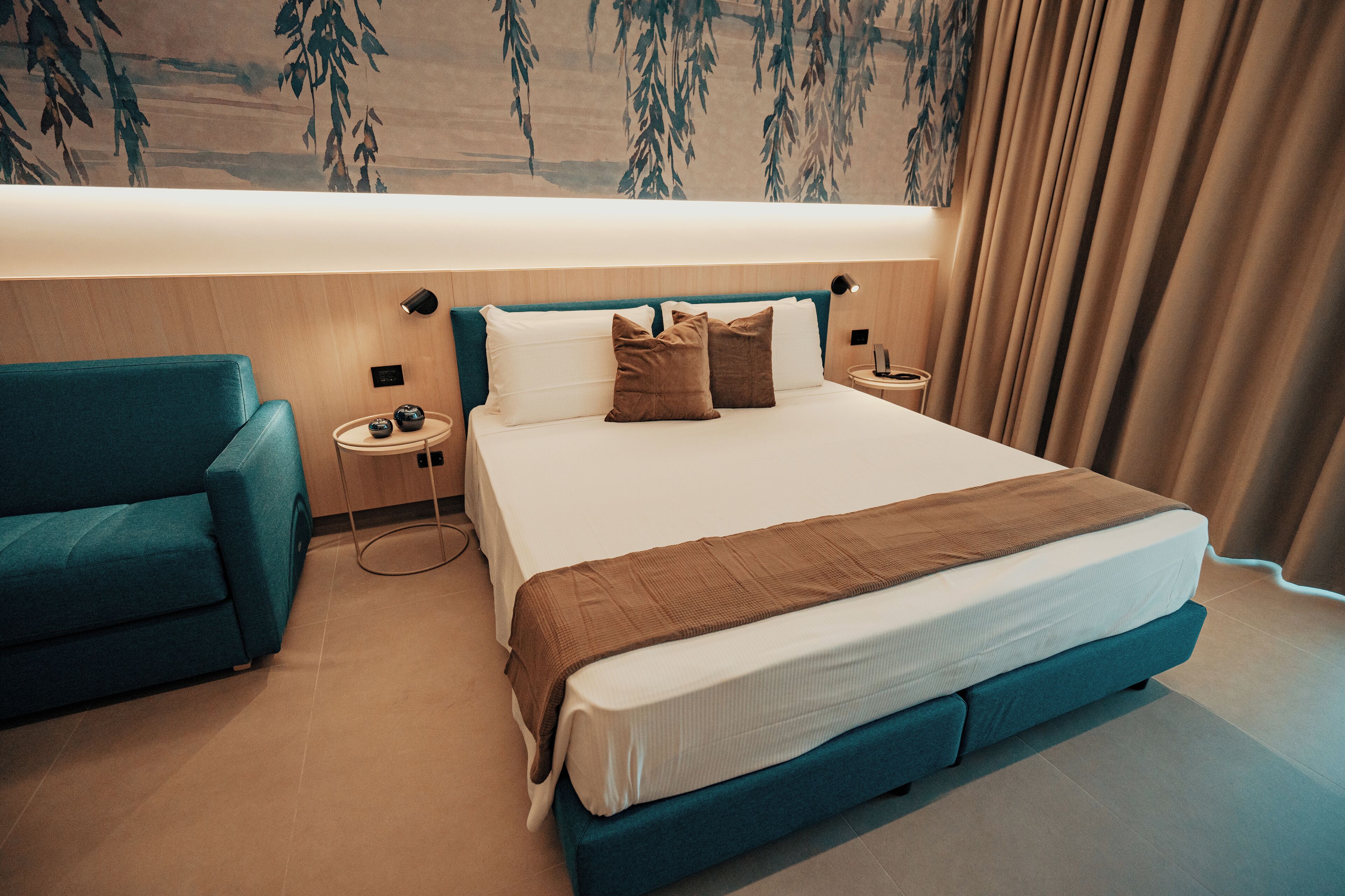 A modern hotel room with a large bed, a teal armchair, a bedside table with a lamp, and a wall mural.