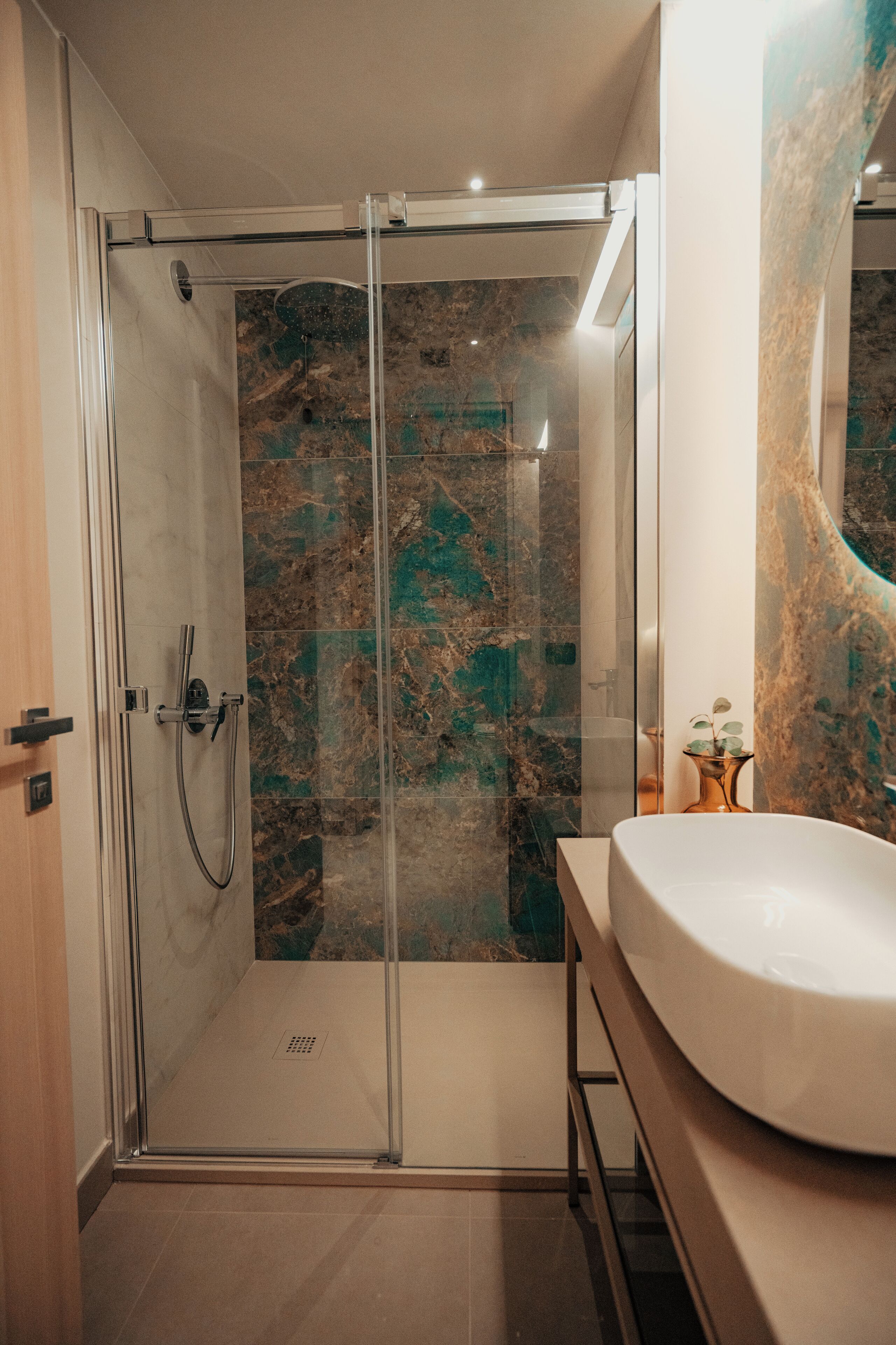 Un baño moderno con ducha envuelta en vidrio, pared con azulejos de mármol y lavamanos blanco.