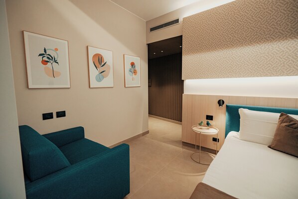 Habitación de hotel moderna con un sofá azul verdoso, cama con cabecera azul y tres cuadros abstractos en la pared.