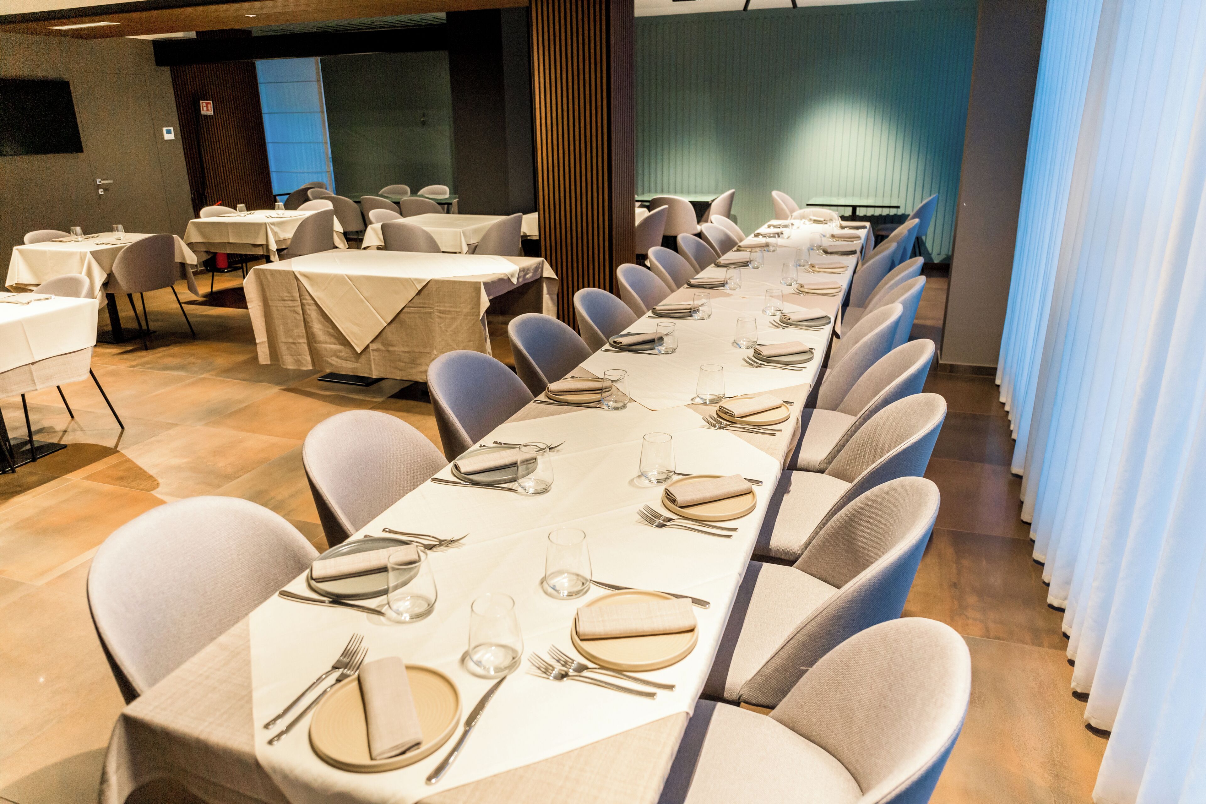 Sala de conferências com mesas preparadas para uma reunião, equipada com cadeiras e mesas, e uma grande janela com cortinas.