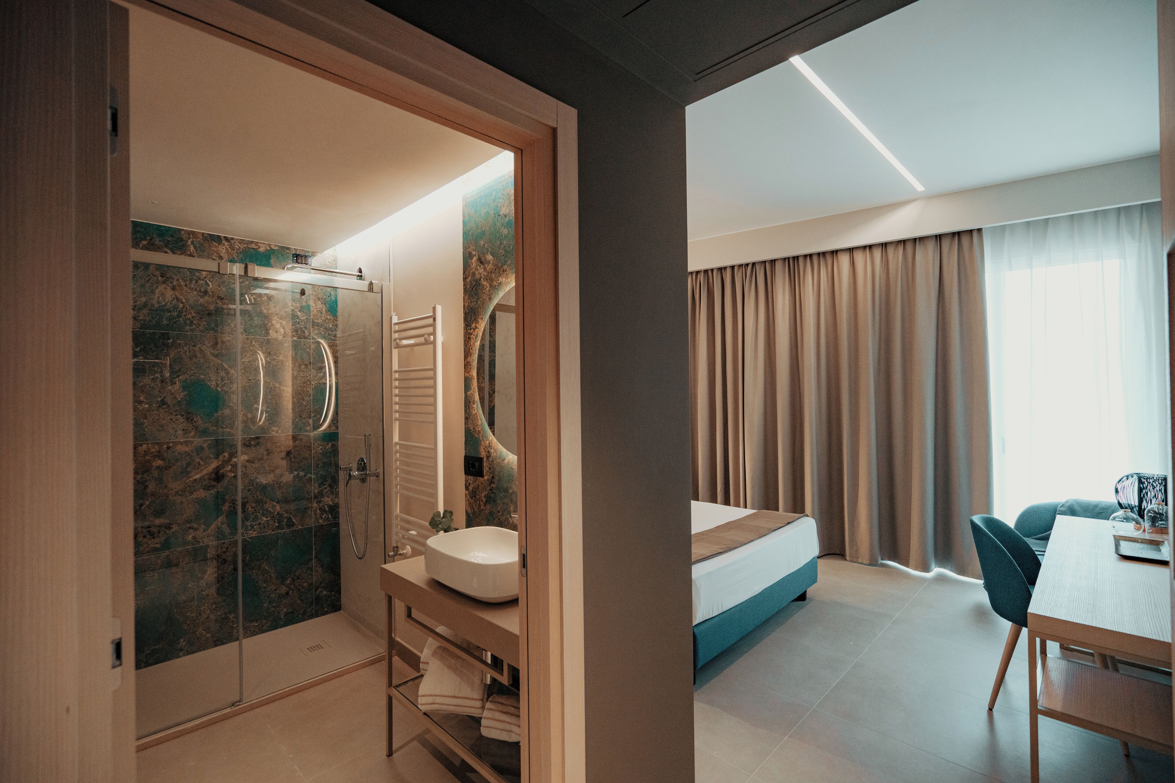 Ein modernes Hotelzimmer mit einer gläsernen Dusche, einem Waschbecken, einem Bett, einem Esstisch und Blick ins Freie.