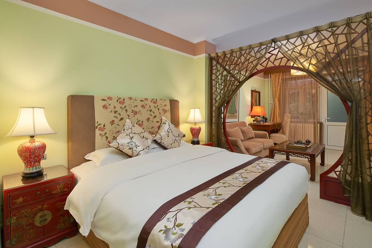 A hotel room with a bed, two bedside lamps, a nightstand, a chair, a coffee table, and a decorative screen.