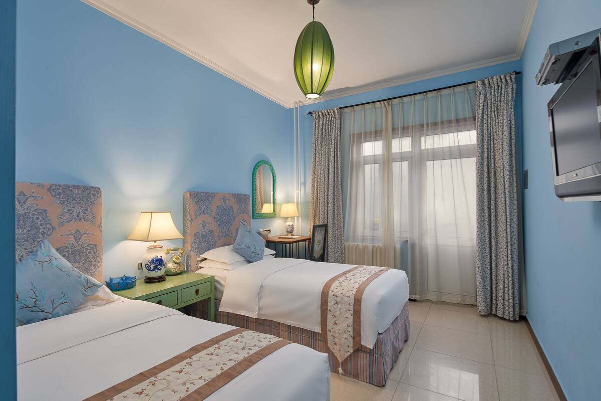 A hotel room with two beds, a green nightstand, a lamp, a mirror, and a large window with curtains.