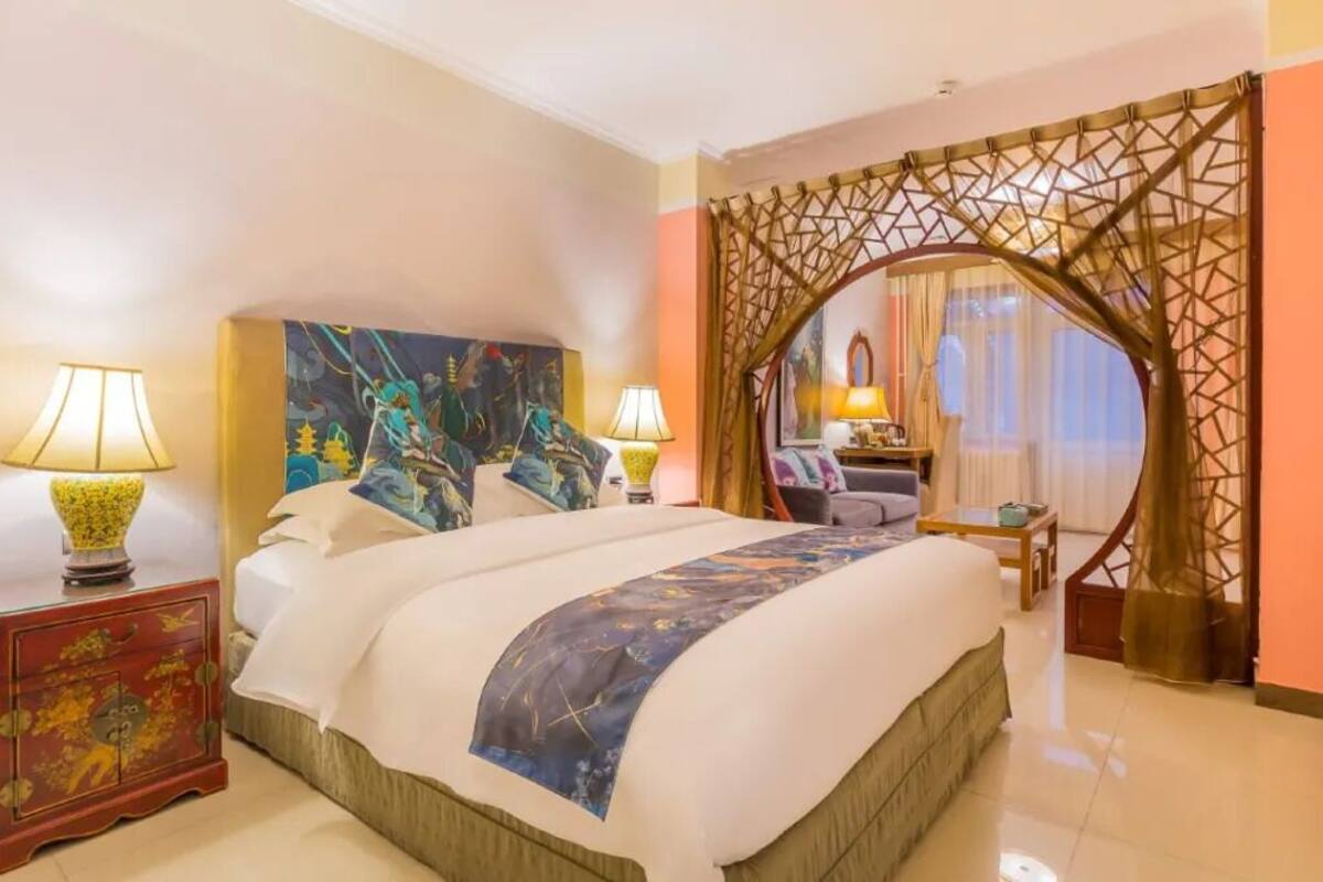 A hotel room with a large bed, two bedside lamps, a decorative headboard, a small table, and a chair.