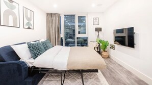 A modern bedroom with a bed, a blue sofa, a TV, and a large window with curtains. - Charming 2BR 2BA near Tower Bridge and Shoreditch (London)