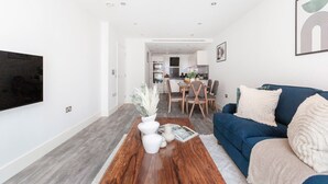 A modern living room with a blue sofa, a coffee table with decorative items, and a flat-screen TV mounted on the wall. - Charming 2BR 2BA near Tower Bridge and Shoreditch (London)