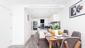 A modern dining area with a wooden table set for six, grey upholstered chairs, and a kitchen in the background. - Charming 2BR 2BA near Tower Bridge and Shoreditch (London)