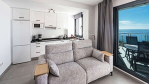 A modern living room with a grey sofa, a wooden side table, and a large window offering a view of the ocean.