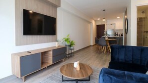 A modern living room with a flat-screen TV mounted on the wall, a dark blue sofa, a wooden coffee table, and a dining area in the background. - w* | Marvelous 2BR with balcony in Miraflores (Lima)