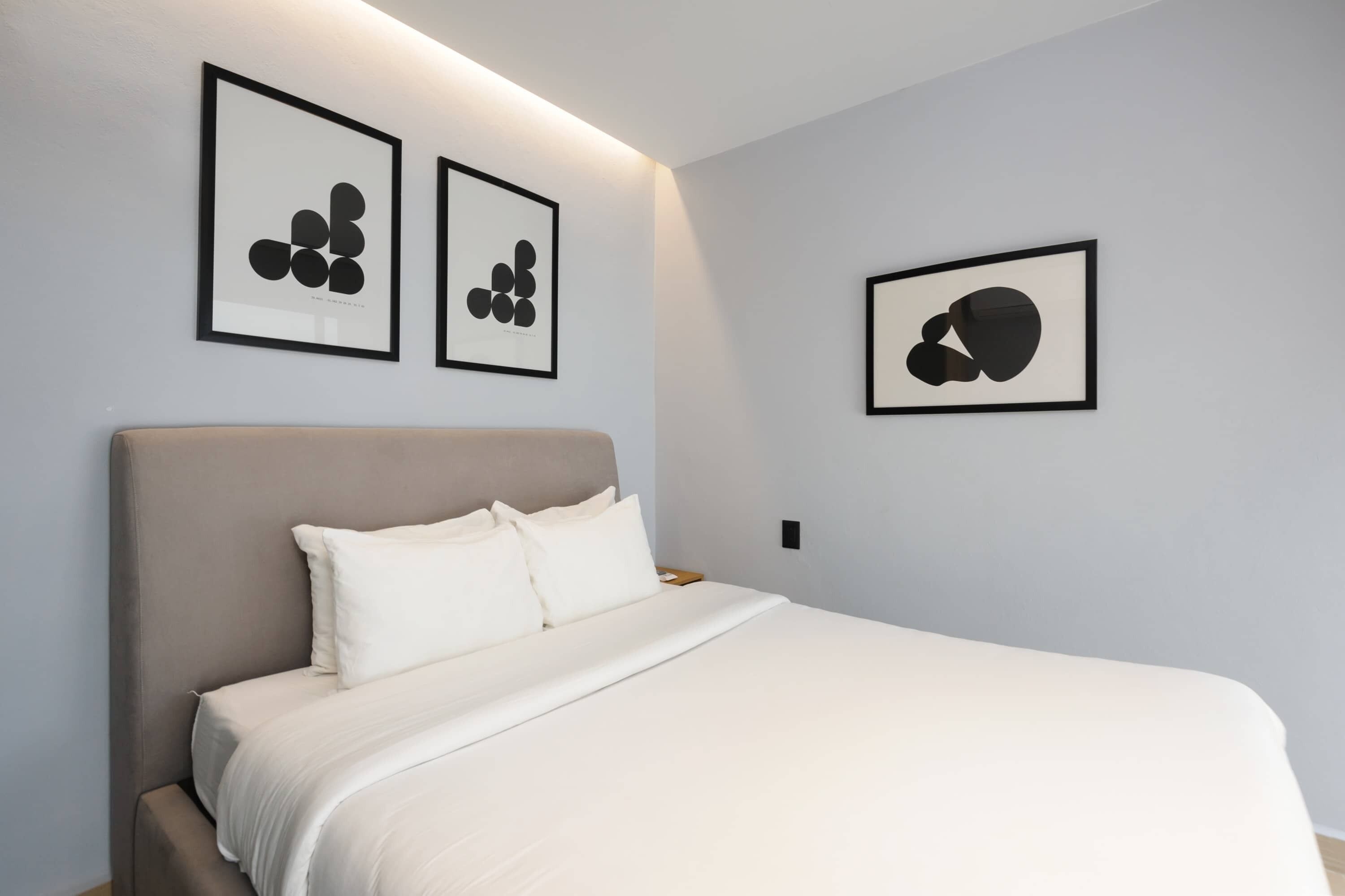 Un lit bien fait, avec du linge de lit blanc et une tête de lit grise, une œuvre d’art encadrée en noir accrochée au mur, et une table de chevet avec une lampe.
