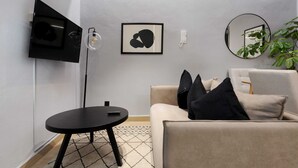 Ein modernes Wohnzimmer mit einer Couch, einem kleinen runden Tisch, einer Stehlampe, einem Flachbildfernseher und gerahmten Kunstwerken an der Wand.