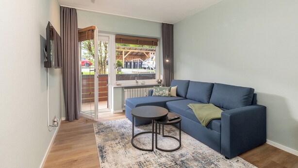 Een woonkamer met een blauwe bank, een klein rond tafeltje en uitzicht op een tuin door een schuifdeur.